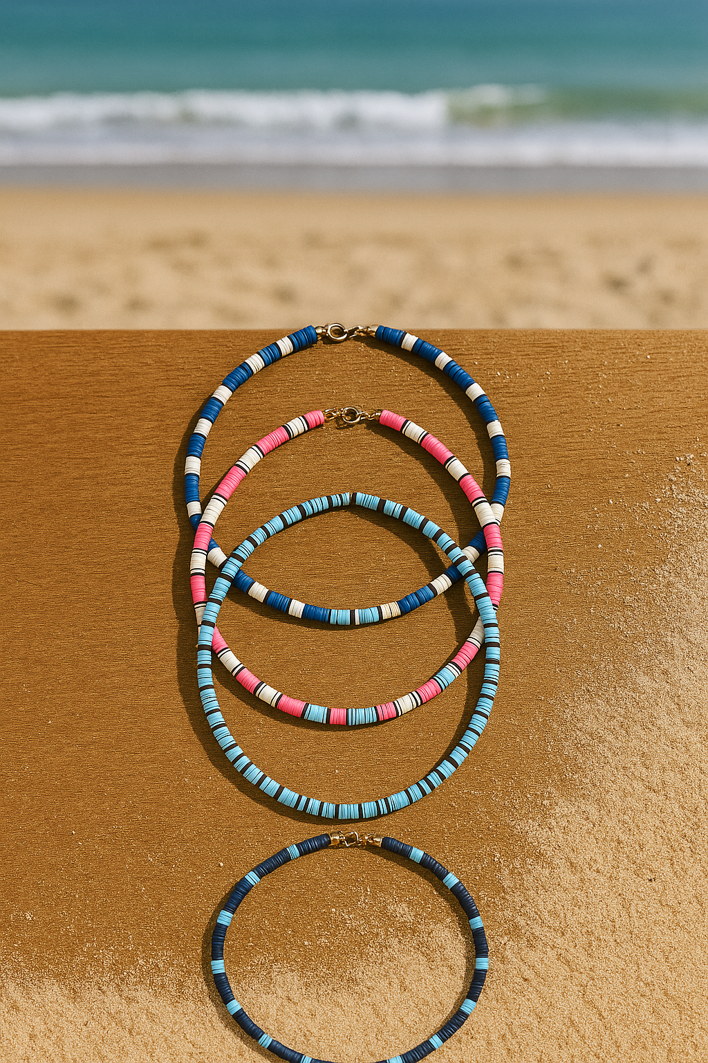 Necklaces on sand with ocean in the background; blue, pink, and turquoise beaded jewelry.