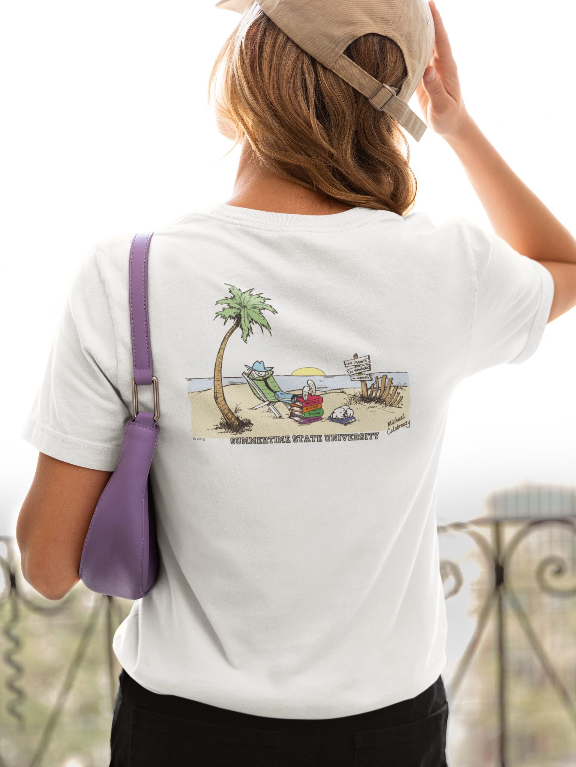 Woman wearing a white t-shirt with a beach scene cartoon on the back. She holds a purple purse and a tan cap.