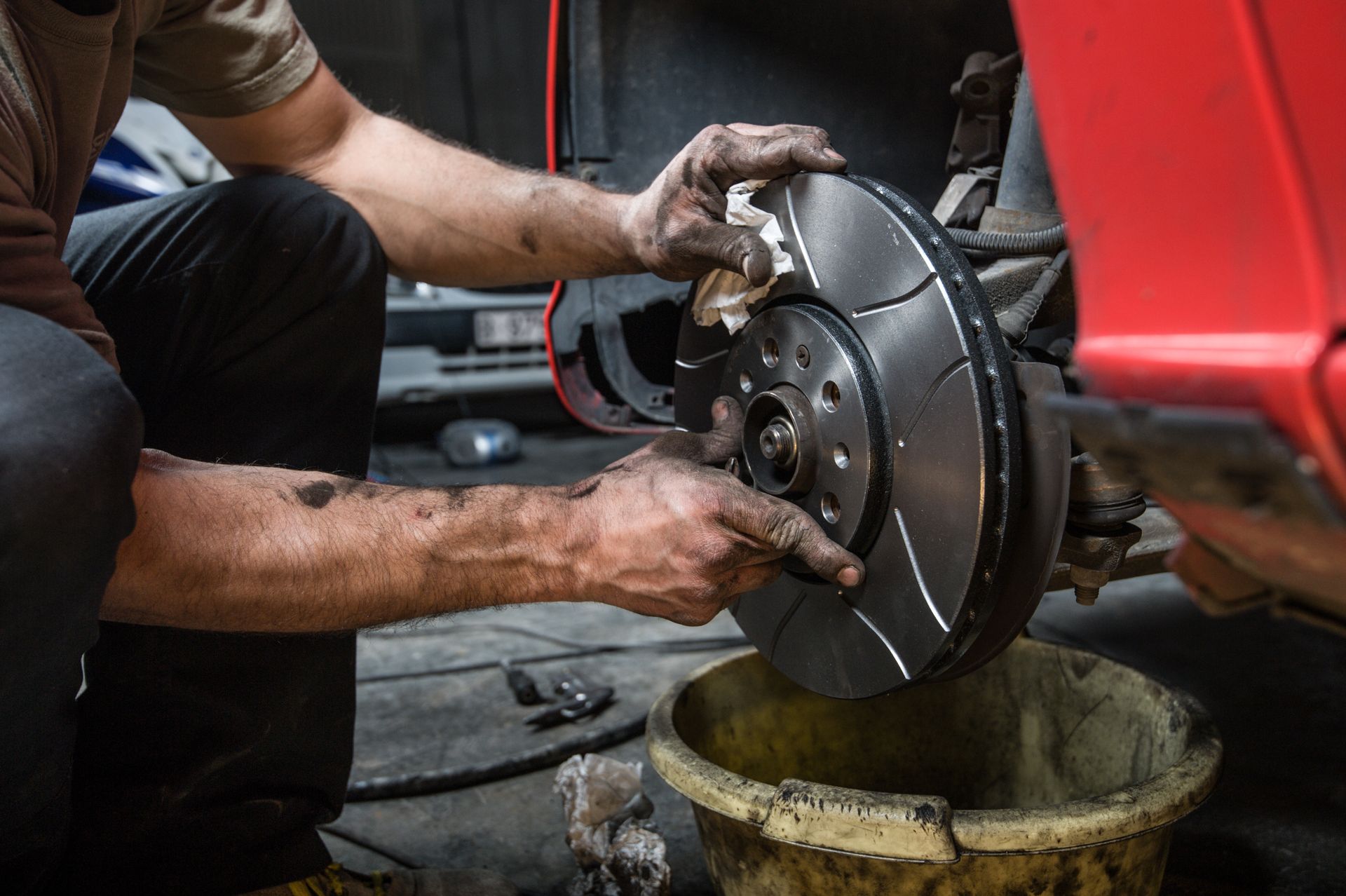 A mechanic with grease-stained hands cleans a ventilated brake rotor on a red car in a garage.