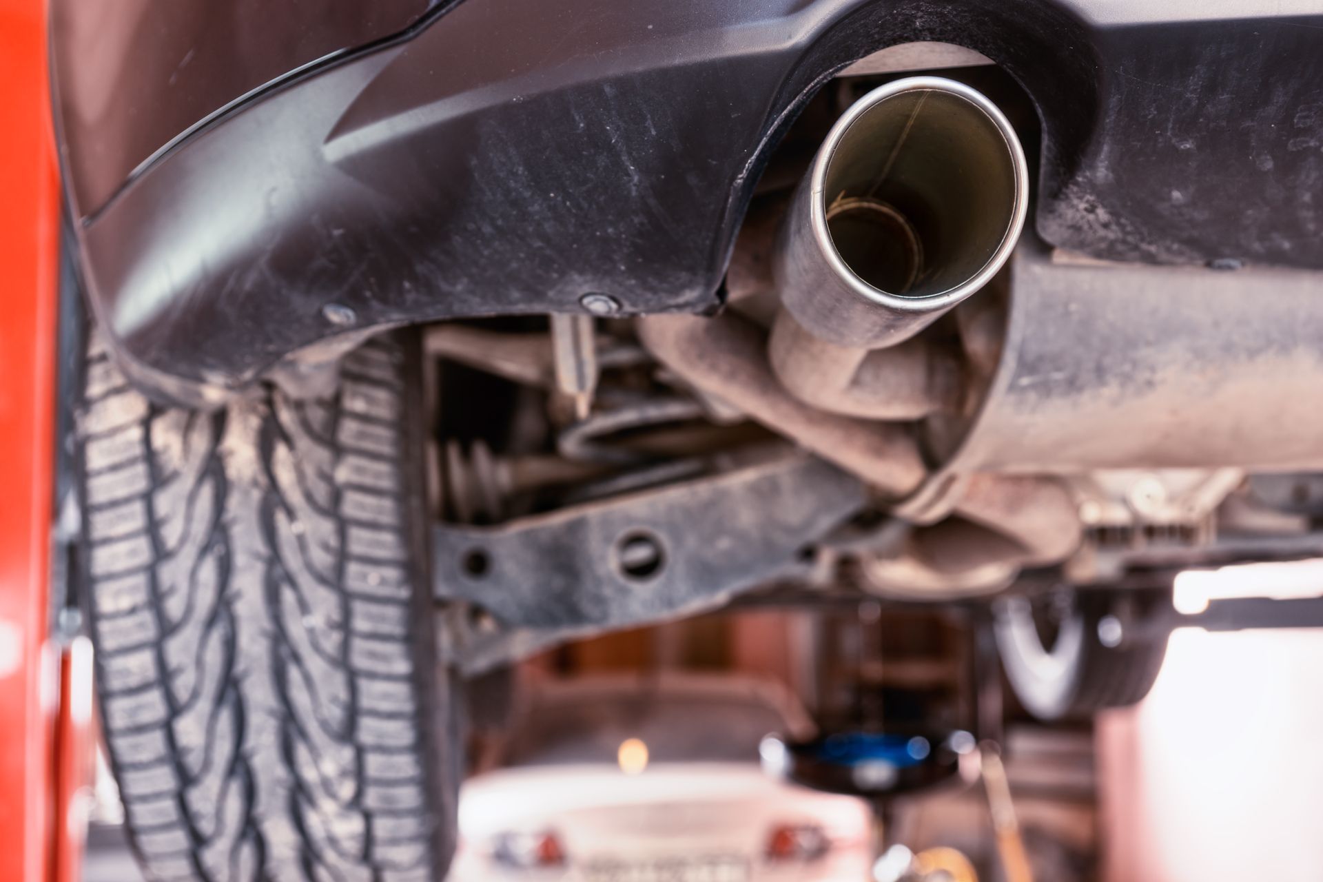 A low-angle view of a car’s undercarriage, highlighting the rear tire, exhaust pipe, and suspension in a garage.