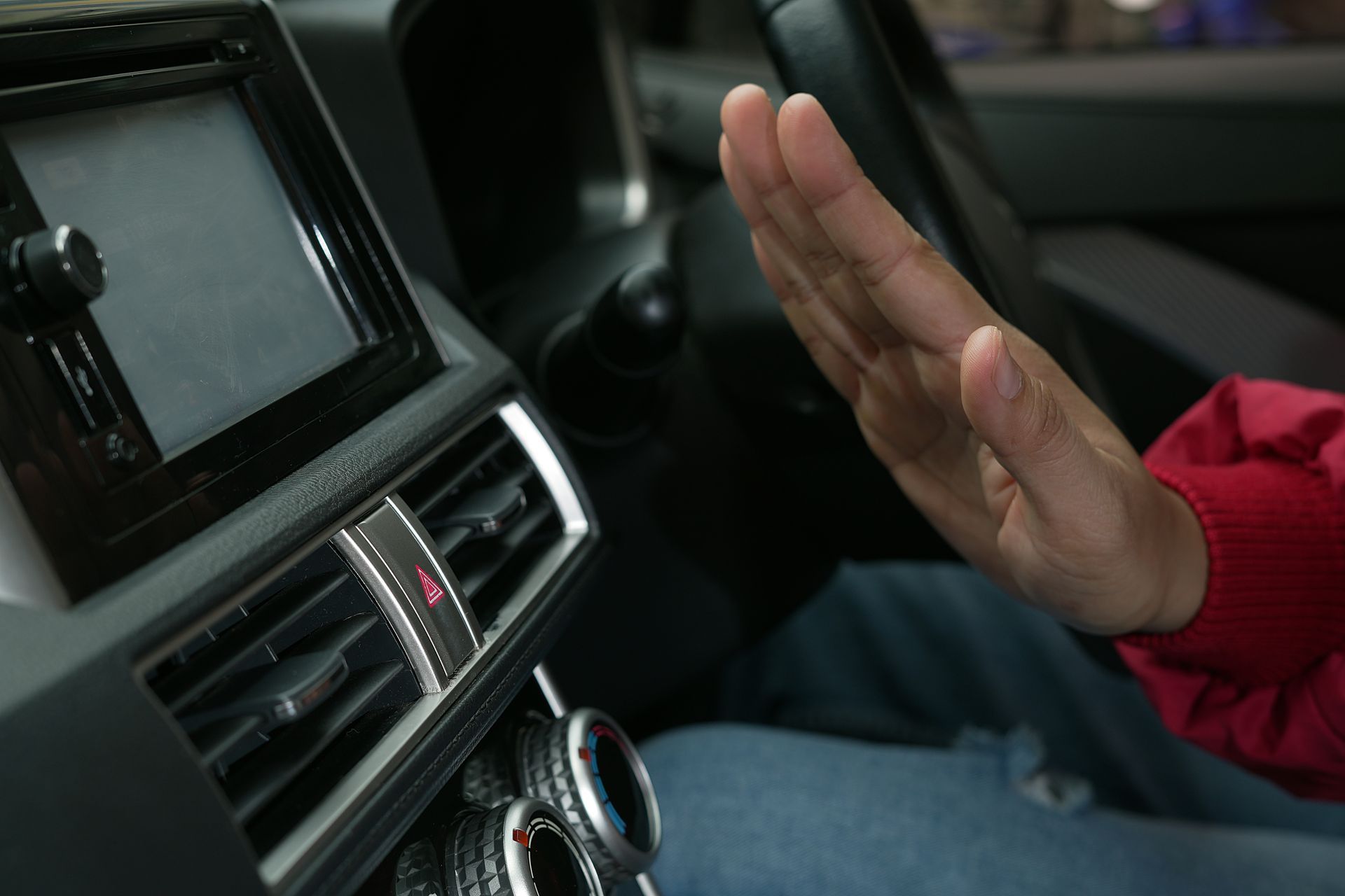 A person in a red sleeve holds their hand up to feel the airflow from a car's dashboard air conditioning vent.