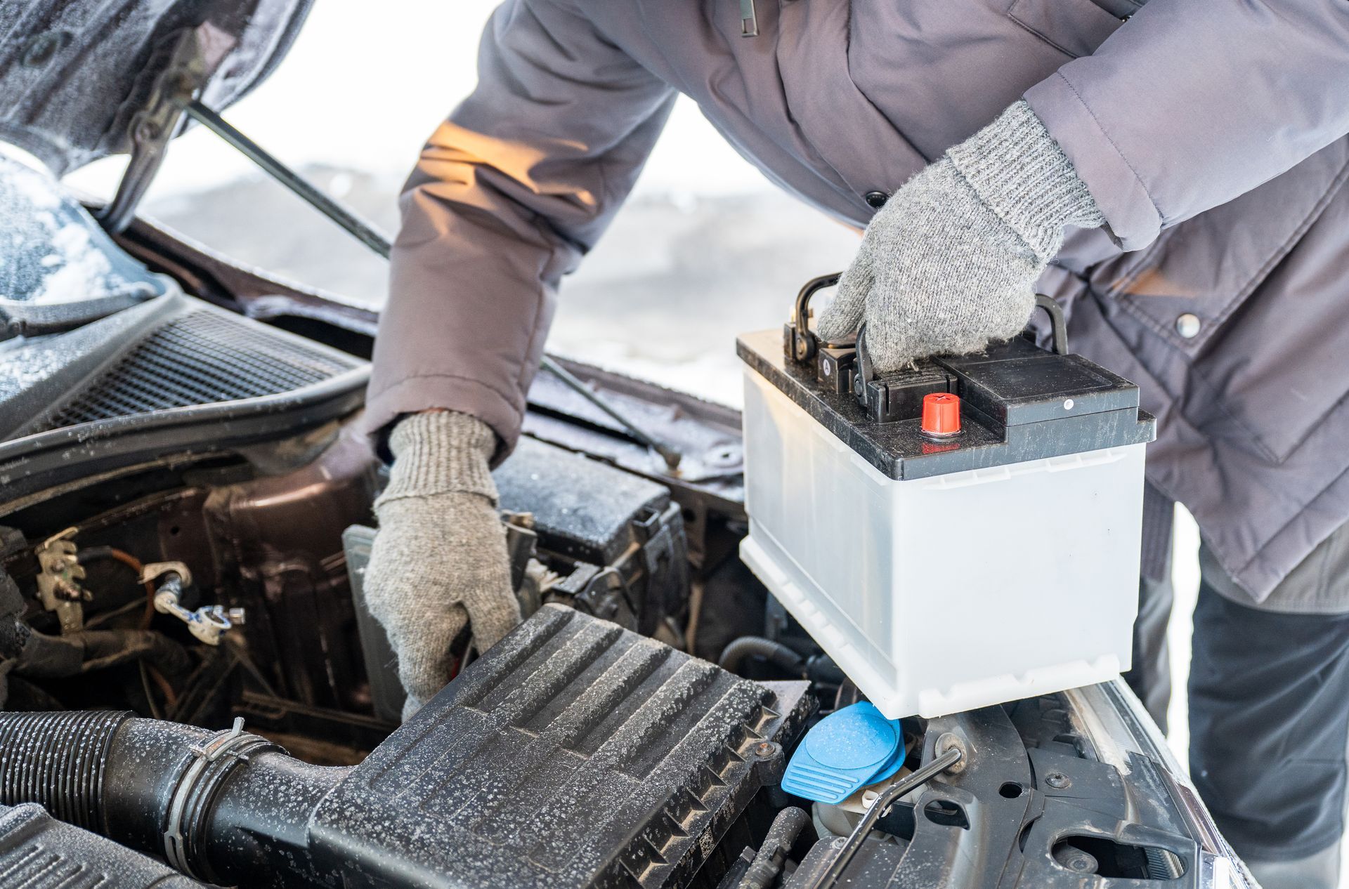 A person wearing winter gloves installs a new car battery into an engine compartment on a snowy day.