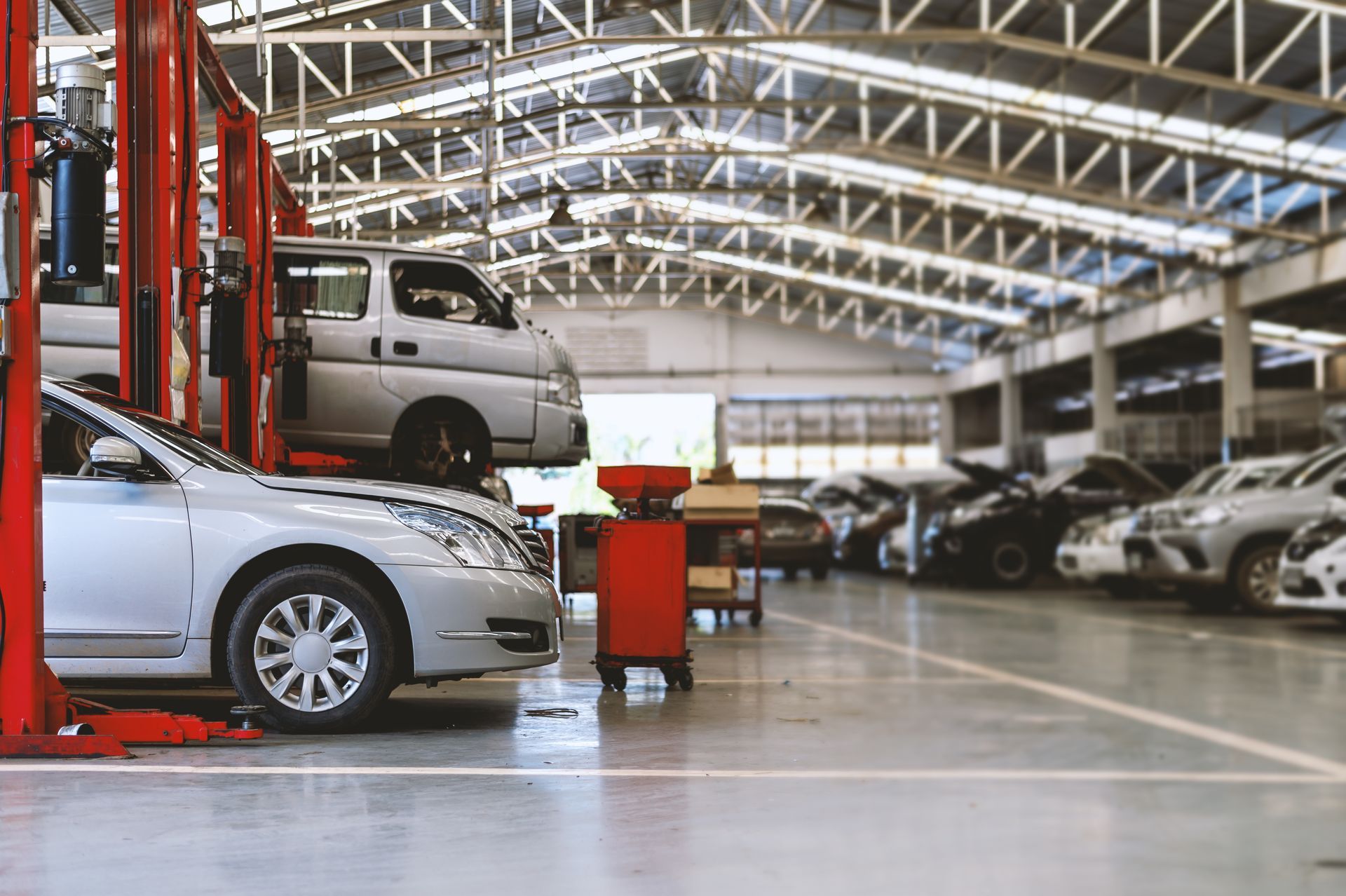 Cars undergoing maintenance inside a spacious, brightly lit auto repair workshop with a high metal-trussed ceiling.