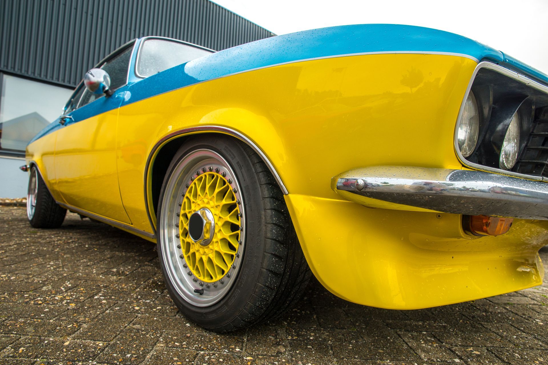 A low-angle view of a yellow and blue vintage car featuring mesh-style wheels parked on a paved lot.