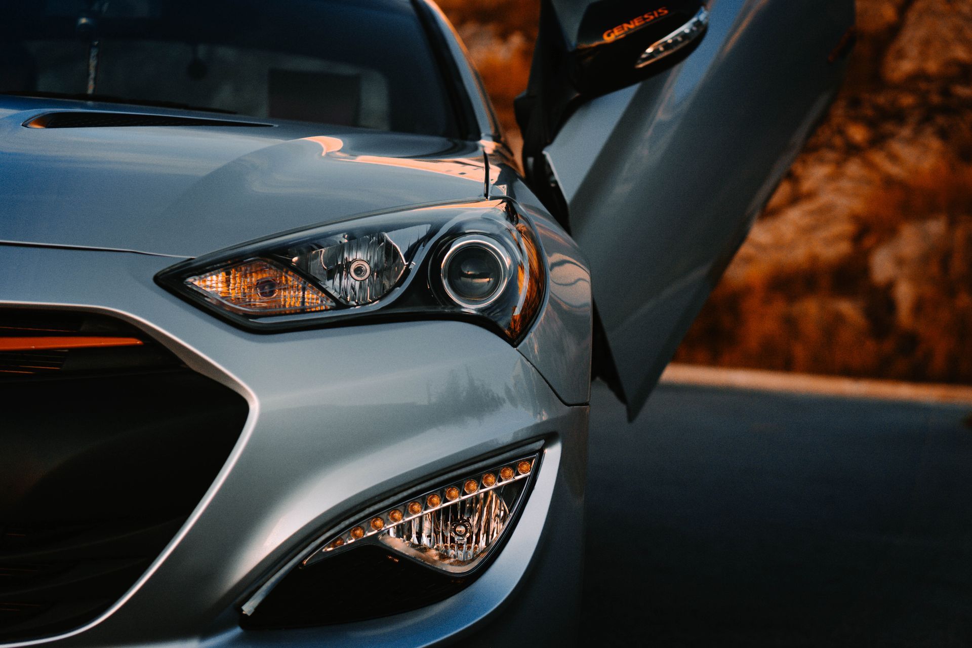 Close-up of a silver sports car with an open scissor door, illuminated by warm, golden-hour light.