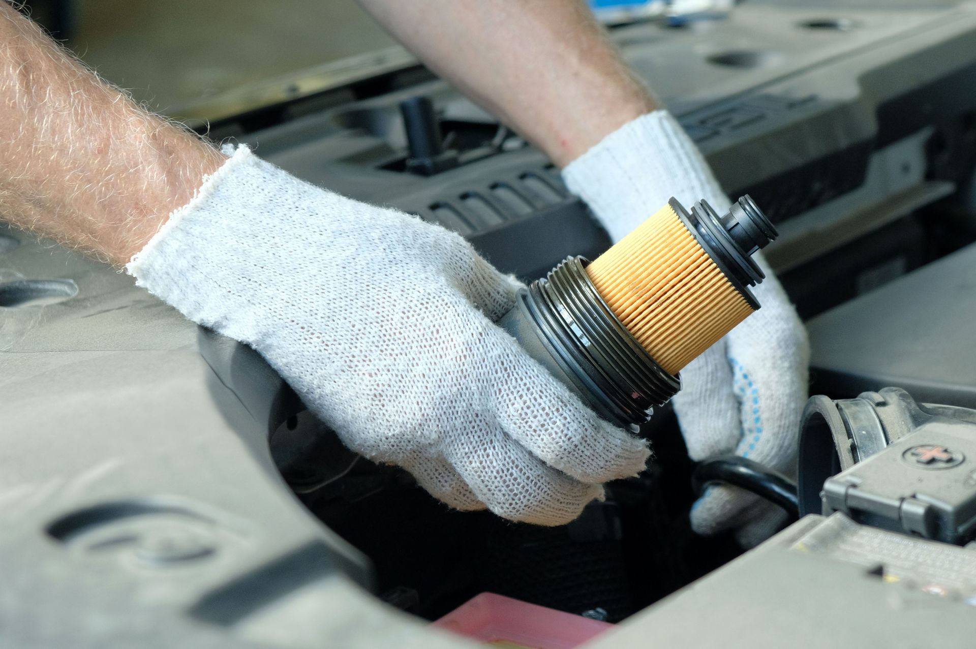 A technician in white work gloves holding a cylindrical car engine oil filter over an open hood.