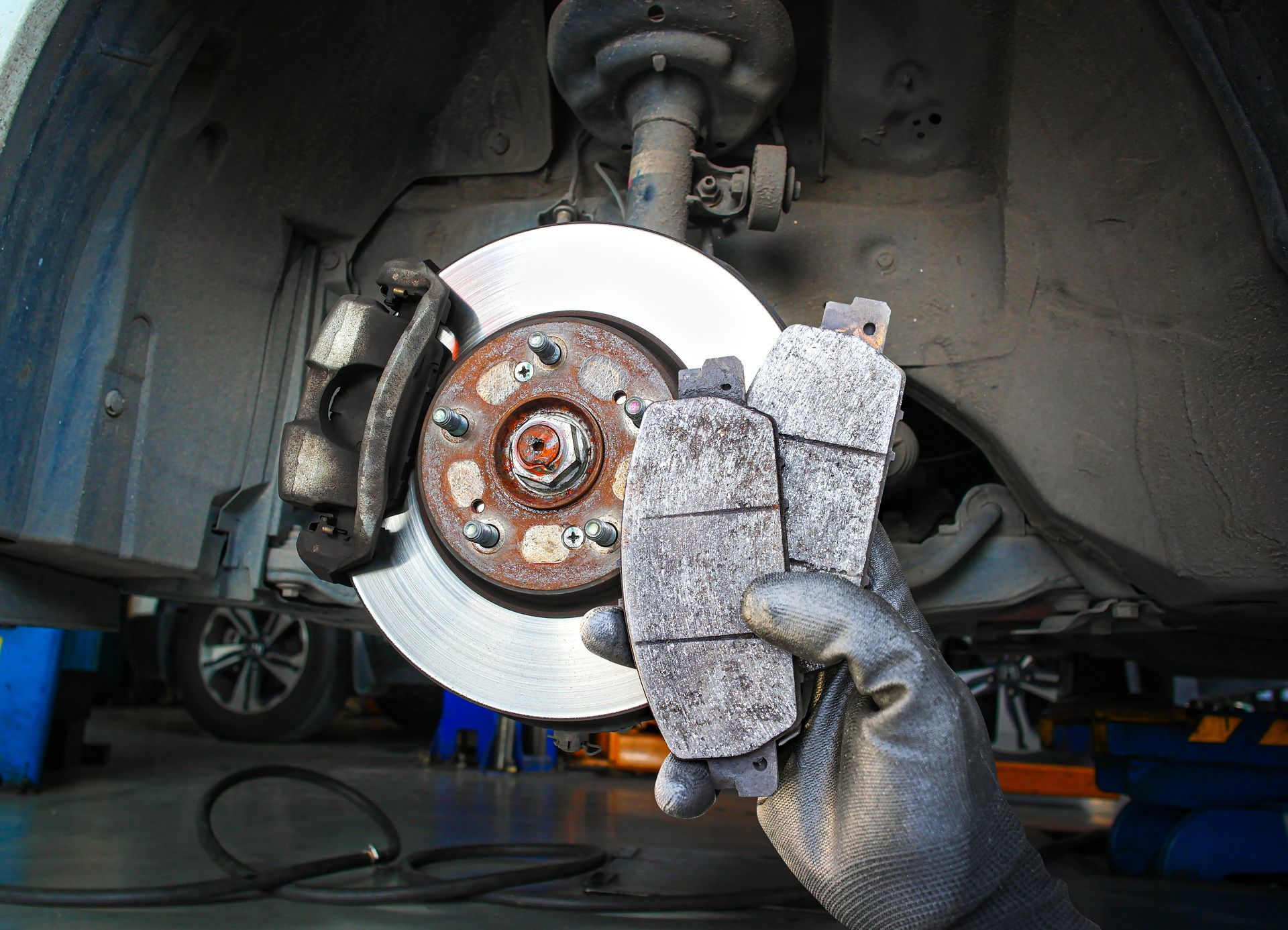 A gloved hand holds worn brake pads in front of a car's exposed wheel assembly and disc brake rotor.
