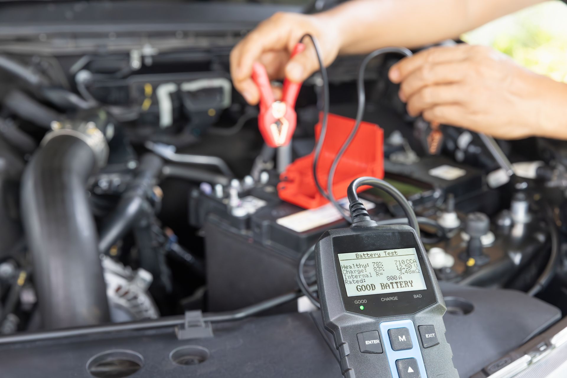 A person testing a car battery using a handheld digital multimeter with red and black clamps attached to the terminals.