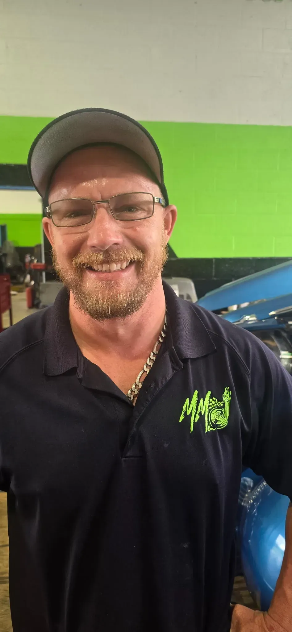 A smiling person wearing glasses, a cap, and a navy polo shirt with a green logo, standing in a workshop.