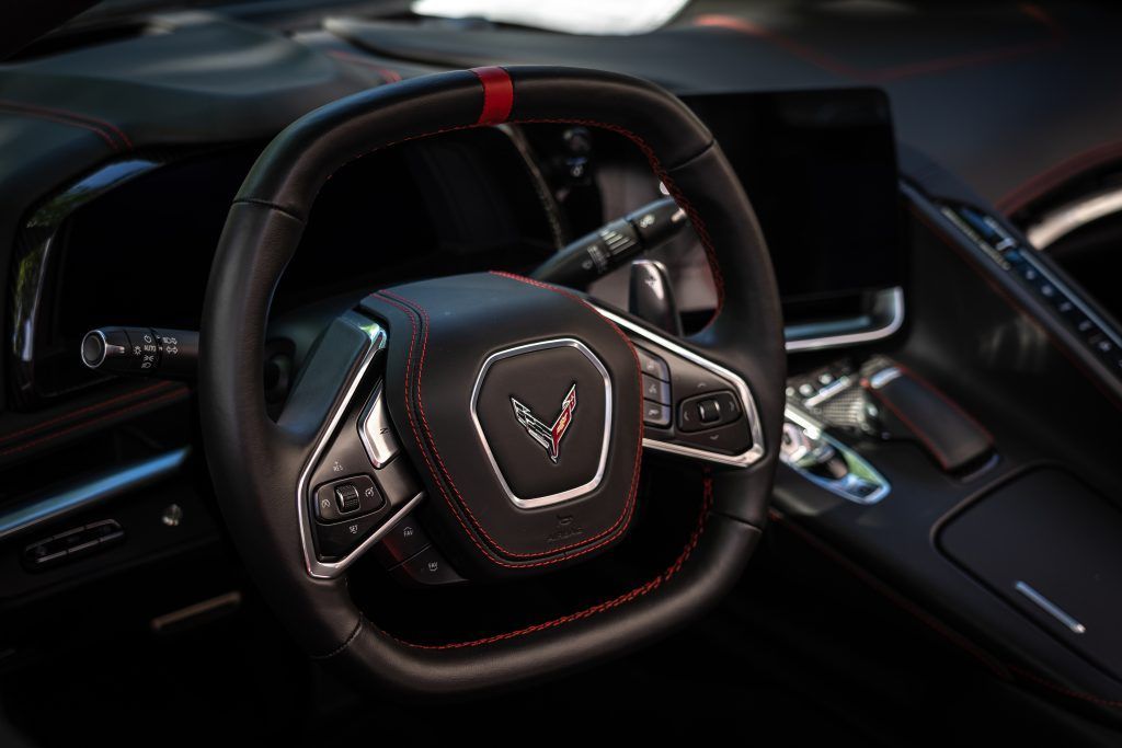 The black leather steering wheel of a Corvette with red stitching and a red racing stripe center mark.