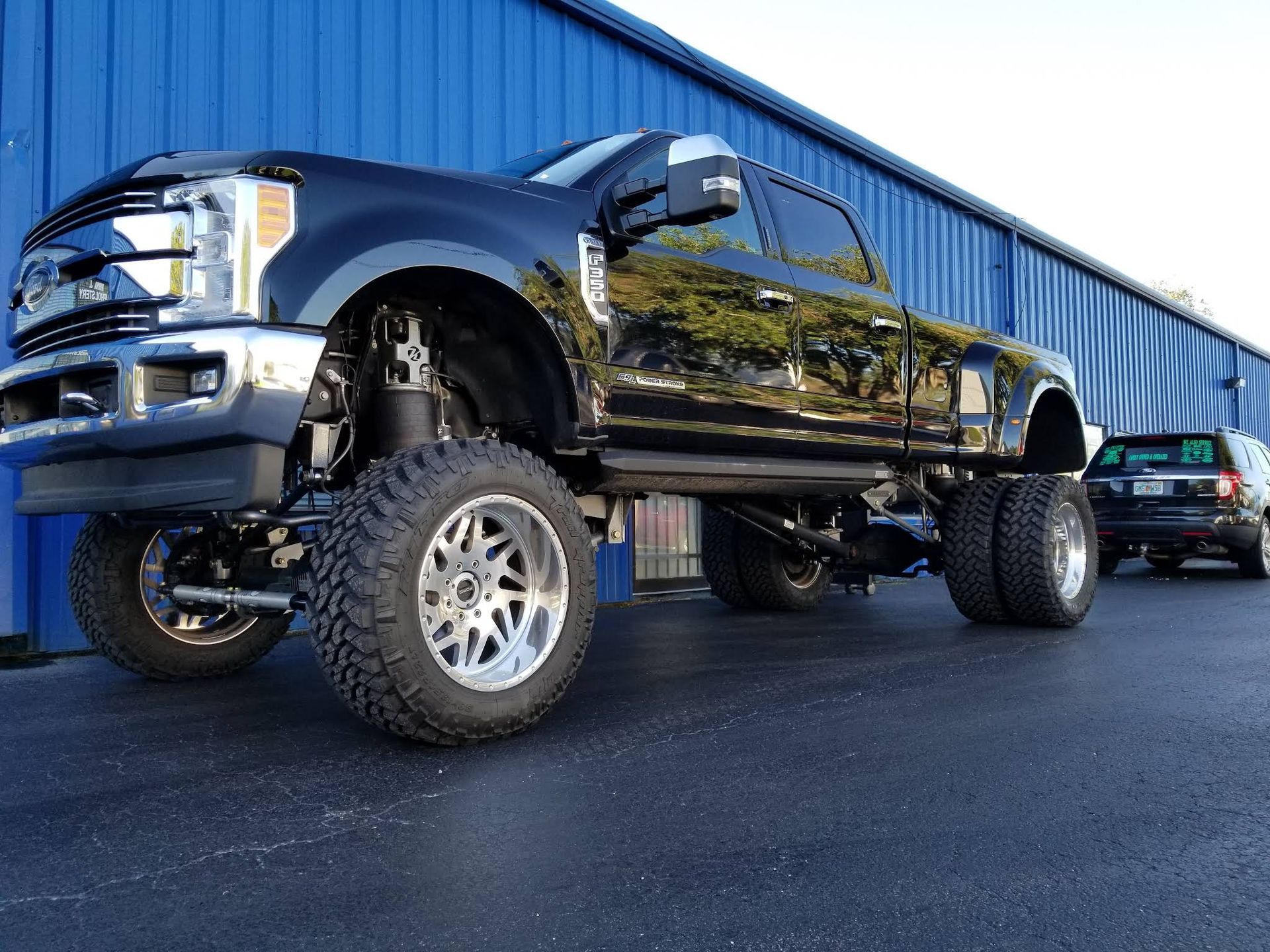 A black lifted dually pickup truck parked on an asphalt lot in front of a blue corrugated metal building.