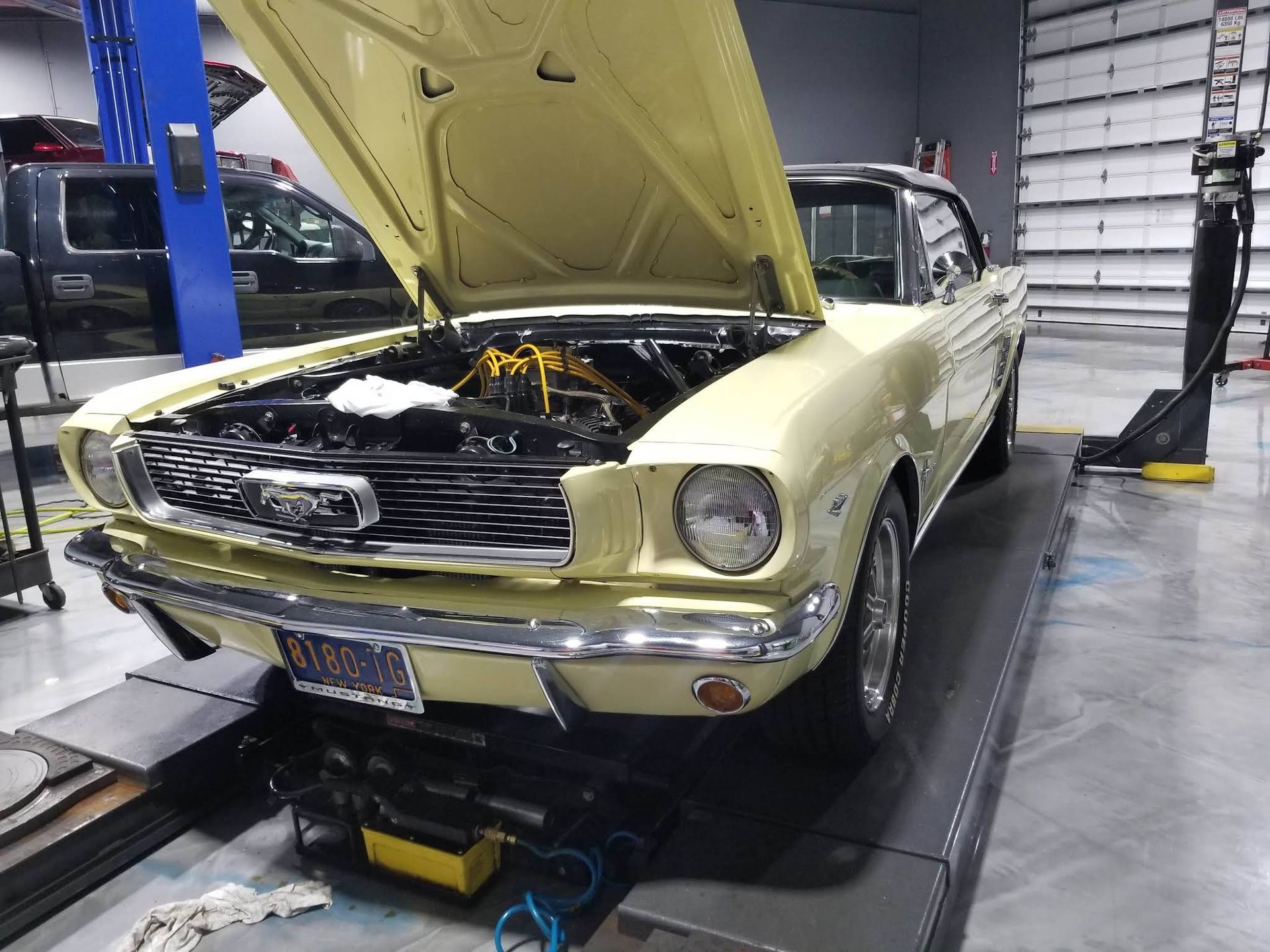 A pale yellow classic Ford Mustang with its hood open, parked on a service lift inside an automotive repair shop.