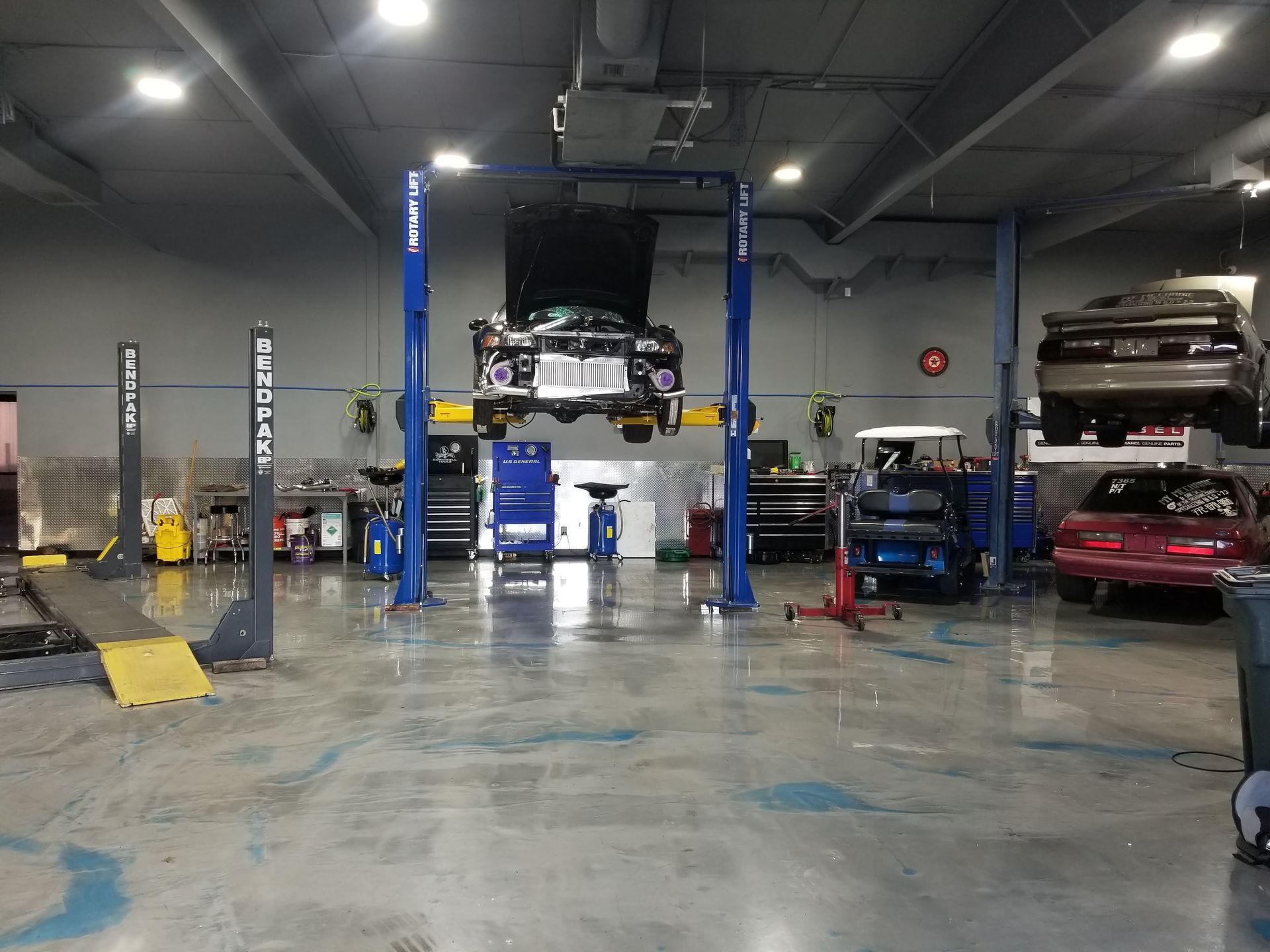 An automotive repair shop with a car on a lift, a utility cart, and other vehicles parked in the background.