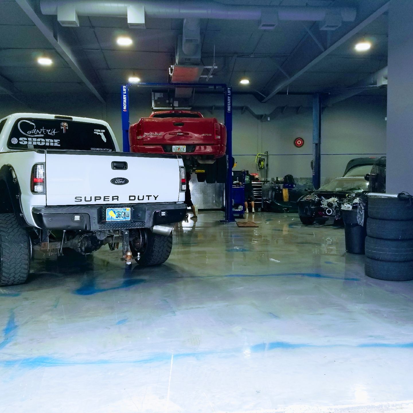 A white Ford Super Duty truck parked in an auto repair shop, with a red truck on a lift and tires stacked to the right.
