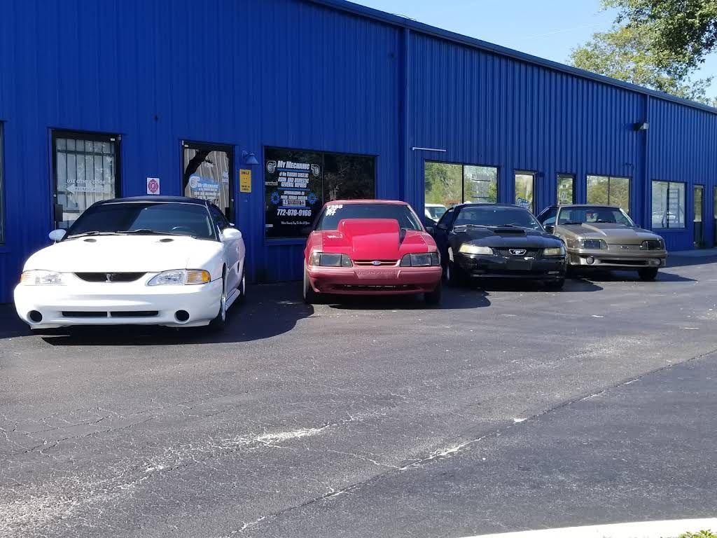 Four Ford Mustangs in white, red, black, and tan are parked in a row in front of a blue industrial building.