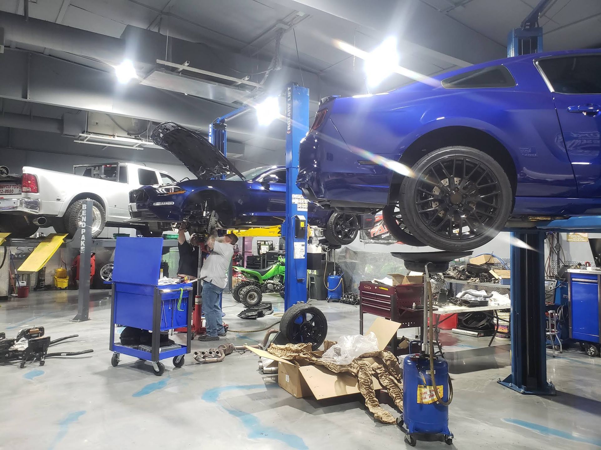 An auto shop with a technician working under a dark blue car lifted on a hoist, near a white truck and tools.