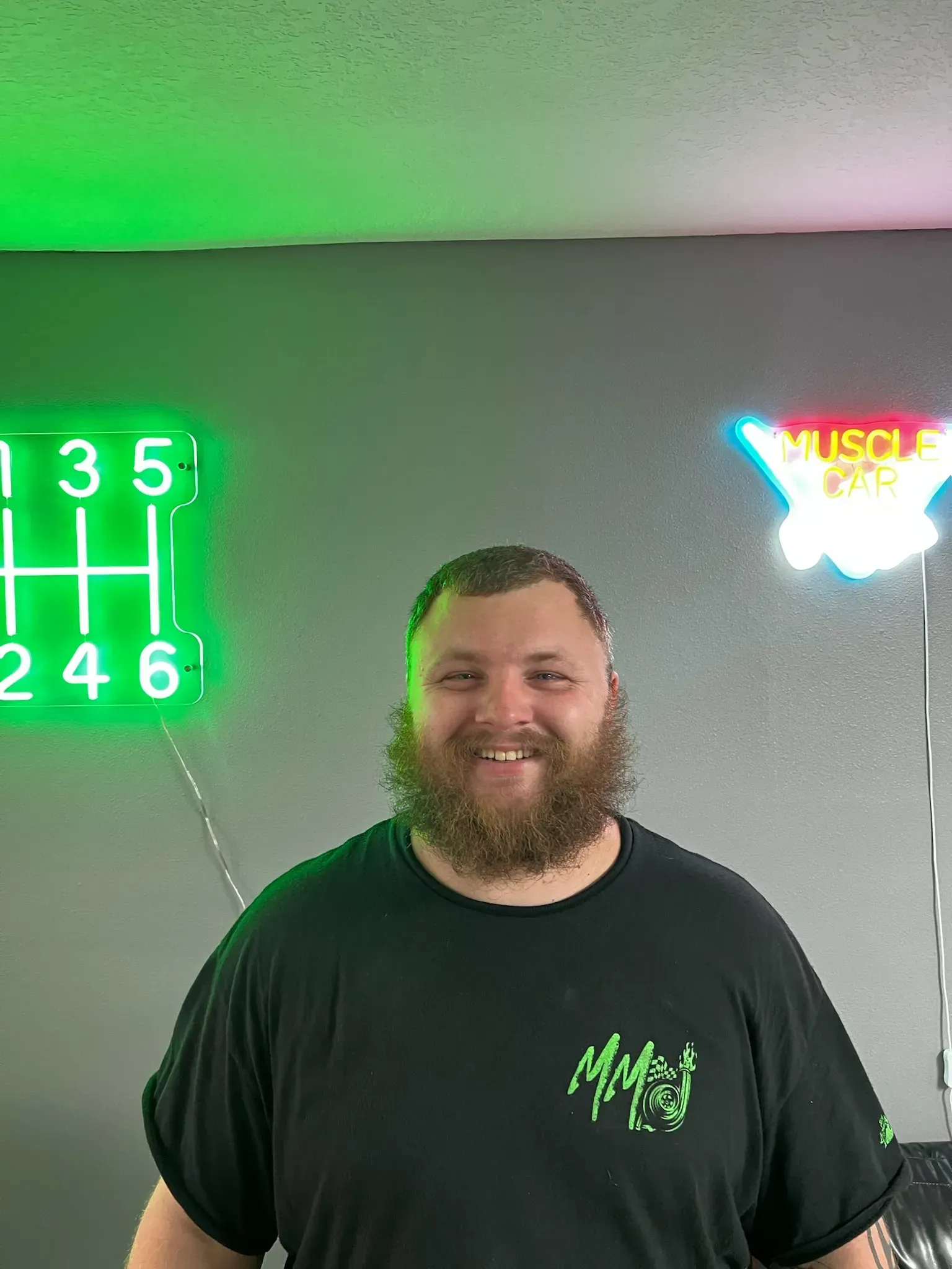 A smiling person with a beard wears a black shirt, standing in front of two neon signs against a gray wall.