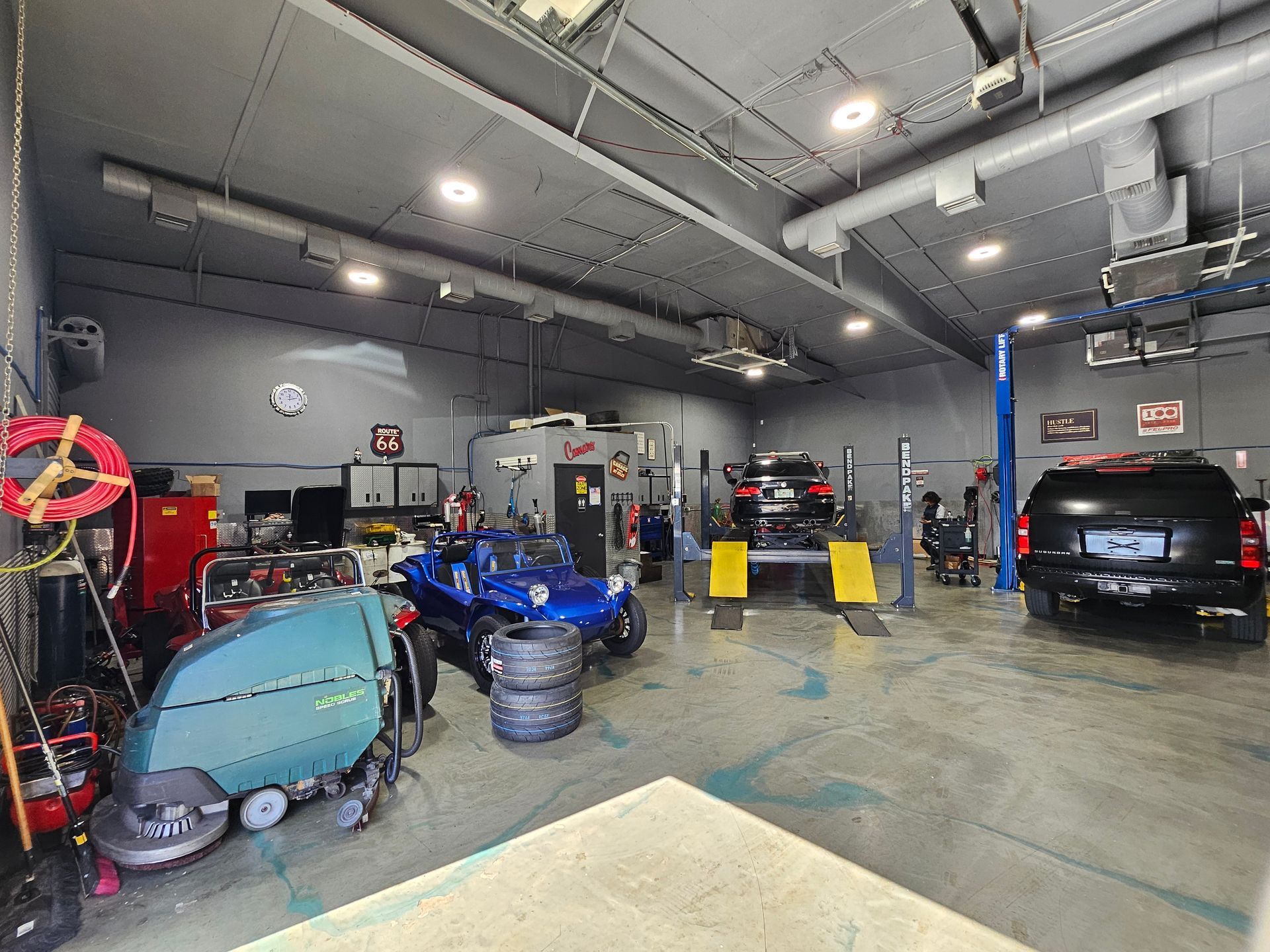 A professional auto repair shop with a black SUV, blue dune buggy, and cleaning equipment on a grey concrete floor.