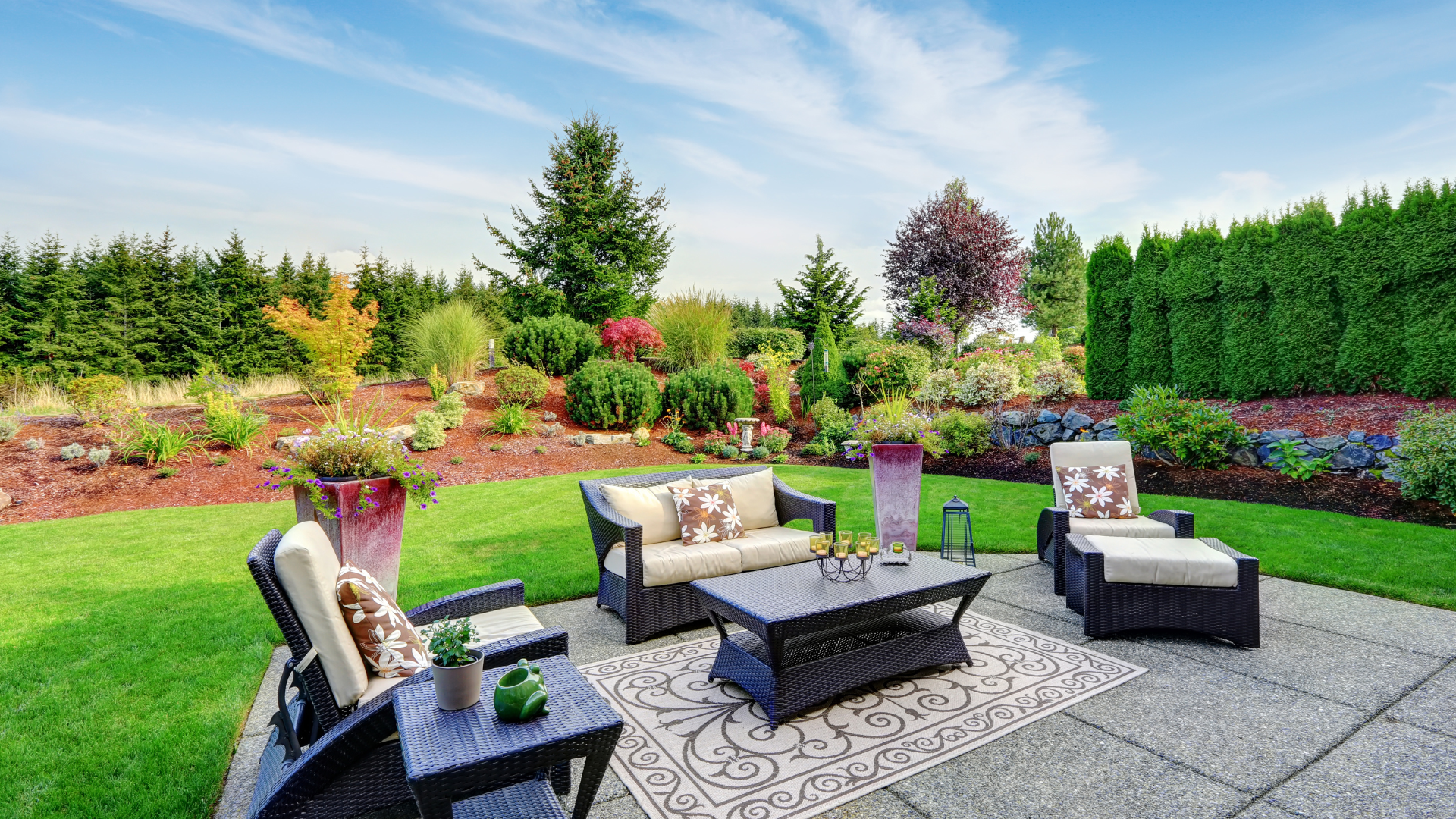 A patio with a couch , chairs , table and rug in a backyard.