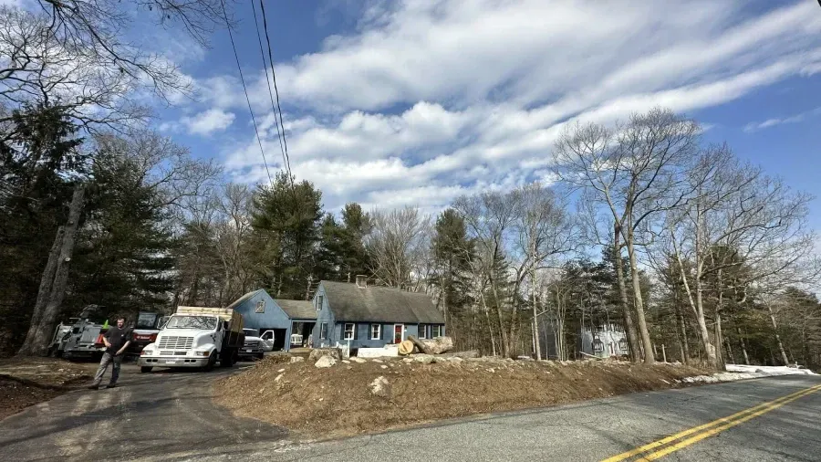 A gray house with a white truck parked in the driveway, situated on a grassy hill next to a road under a partly cloudy sky.