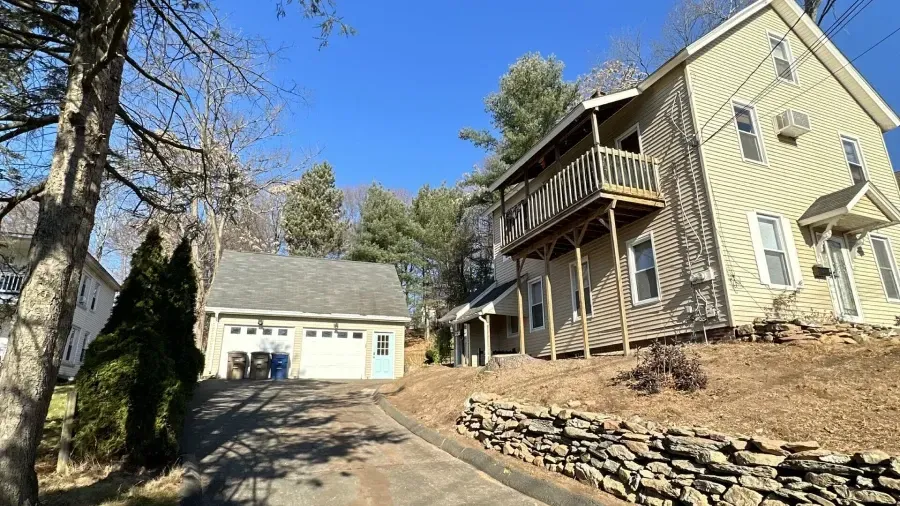 A pale yellow house with a two-story deck, a detached garage, and a stone retaining wall under a clear blue sky.
