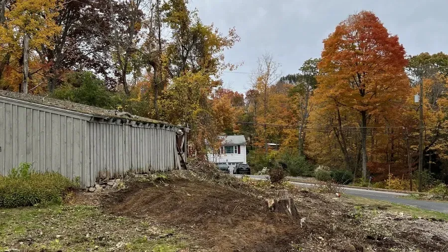 A weathered gray wooden structure sits near a dirt clearing with a small house and vibrant autumn trees in the background.