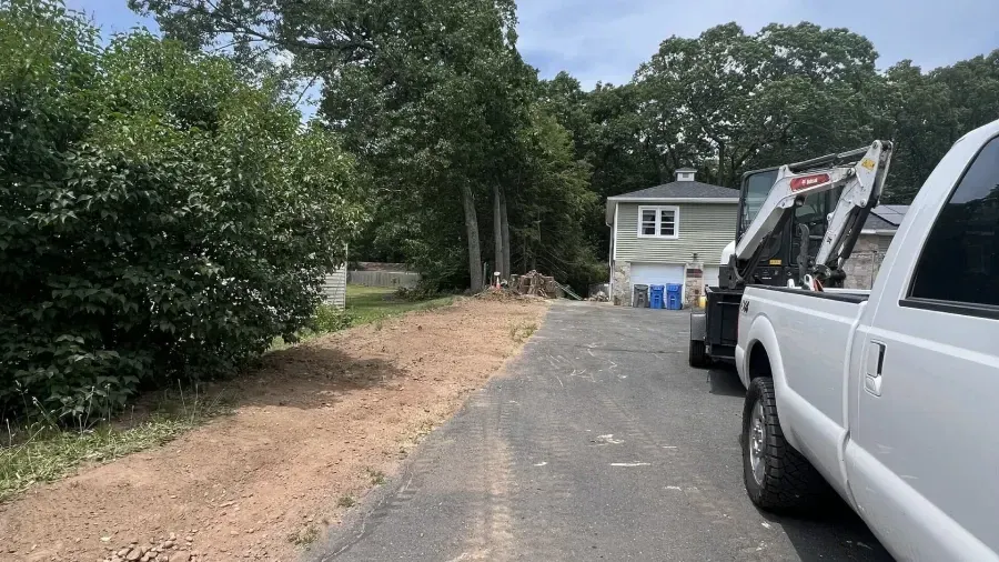 A white pickup truck with a trailer carrying a skid steer parked on a paved driveway next to a cleared dirt patch.