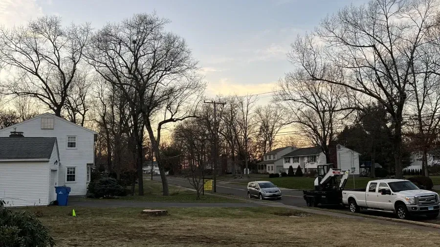 A residential street scene at dusk with a white house on the left, trees, a parked car, and a white pickup truck.
