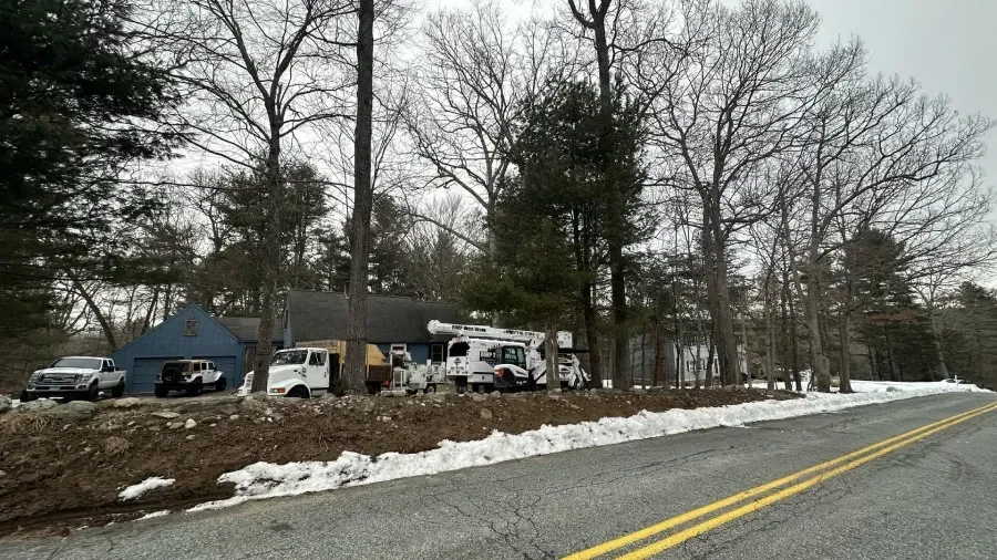 Utility trucks are parked in front of a house beside a wooded road with patches of snow on the ground.