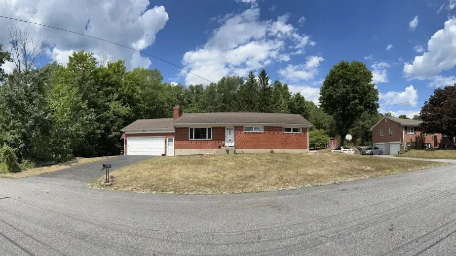 A one-story brick ranch-style house with an attached garage, set on a grassy corner lot under a blue, cloudy sky.