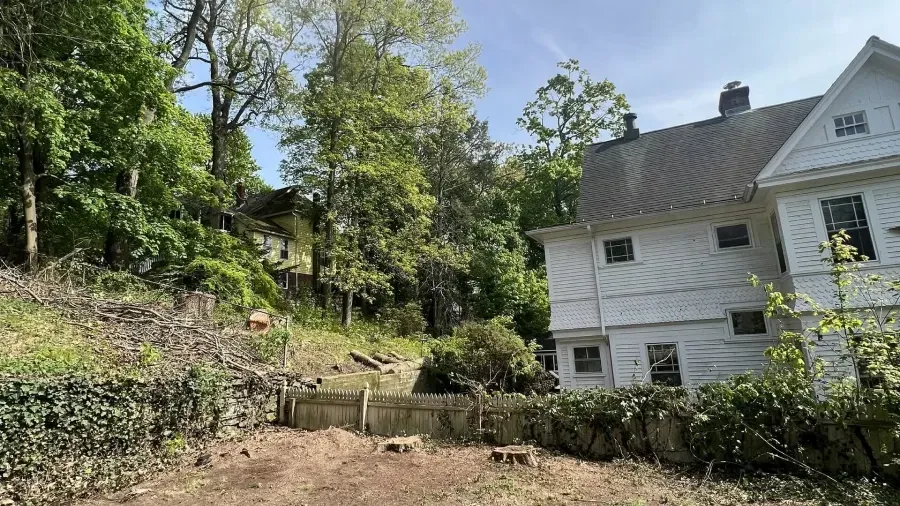 A white house sits next to a wooded hillside with a fence in the foreground.
