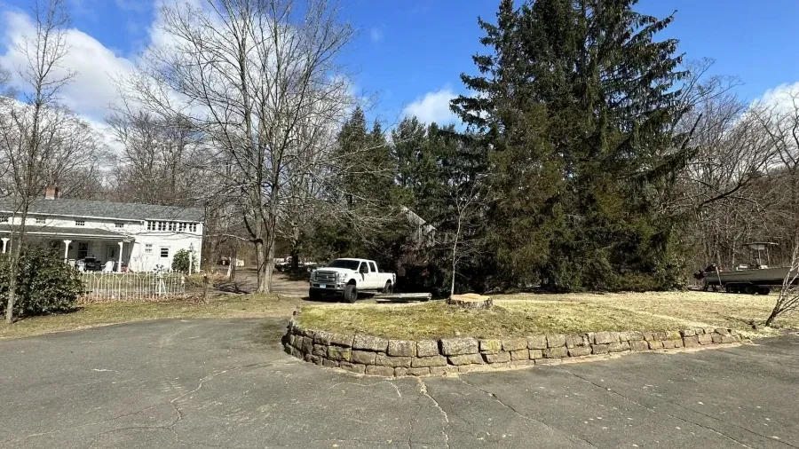 A white pickup truck parked in a driveway in front of a white house with a stone retaining wall and evergreen trees.