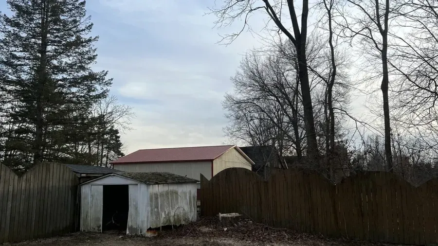 A small, weathered white shed stands in a yard before a fence, with a larger tan building with a red roof behind it.