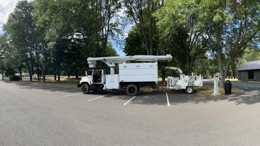 A white utility truck with a bucket lift and a tow-behind wood chipper parked in a parking lot near trees.