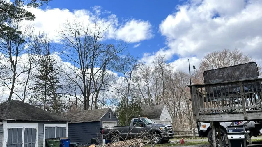 A grey pickup truck parked in a yard with small buildings and a raised wooden deck under a cloudy blue sky.