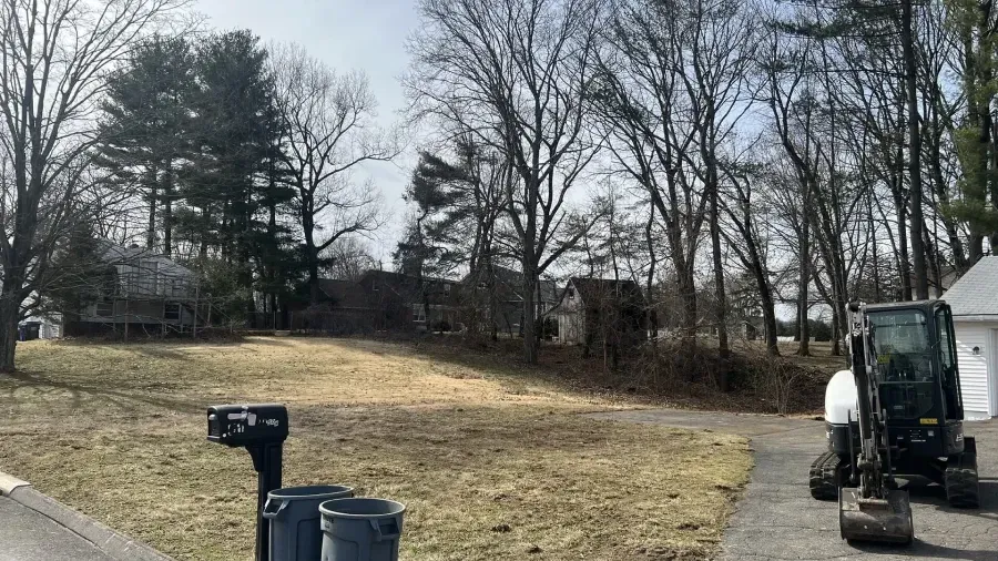 A construction excavator sits on a driveway next to a yard with a mailbox, trash cans, trees, and houses.