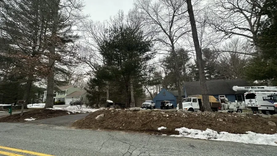 A road view of a home with a large pile of dirt, several vehicles, and trees in a winter landscape.
