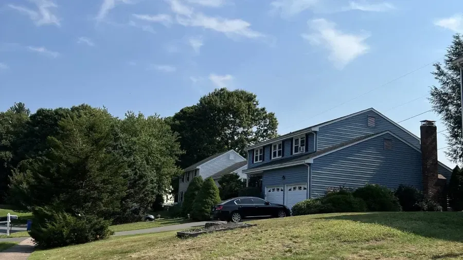 Blue suburban house with a two-car garage and a brick chimney under a clear blue sky, set on a sloped grassy lawn.