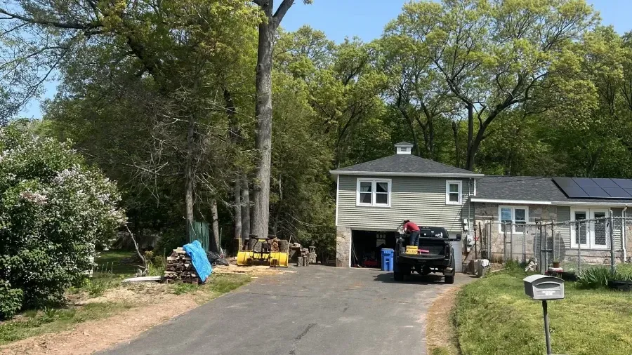 A person stands near a truck parked in a driveway in front of a house with gray siding and lush trees in the background.