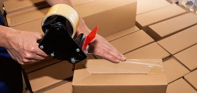 Person sealing a cardboard box with packing tape. Boxes are stacked in the background.