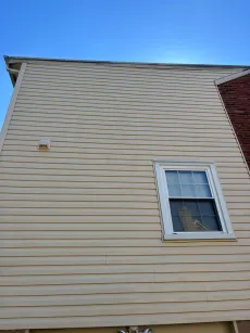 Tan siding on a house with a window and a brick wall section, under a blue sky.