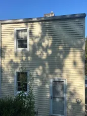Side of a two-story beige house with a window, door, and a bird on the roof under a blue sky.
