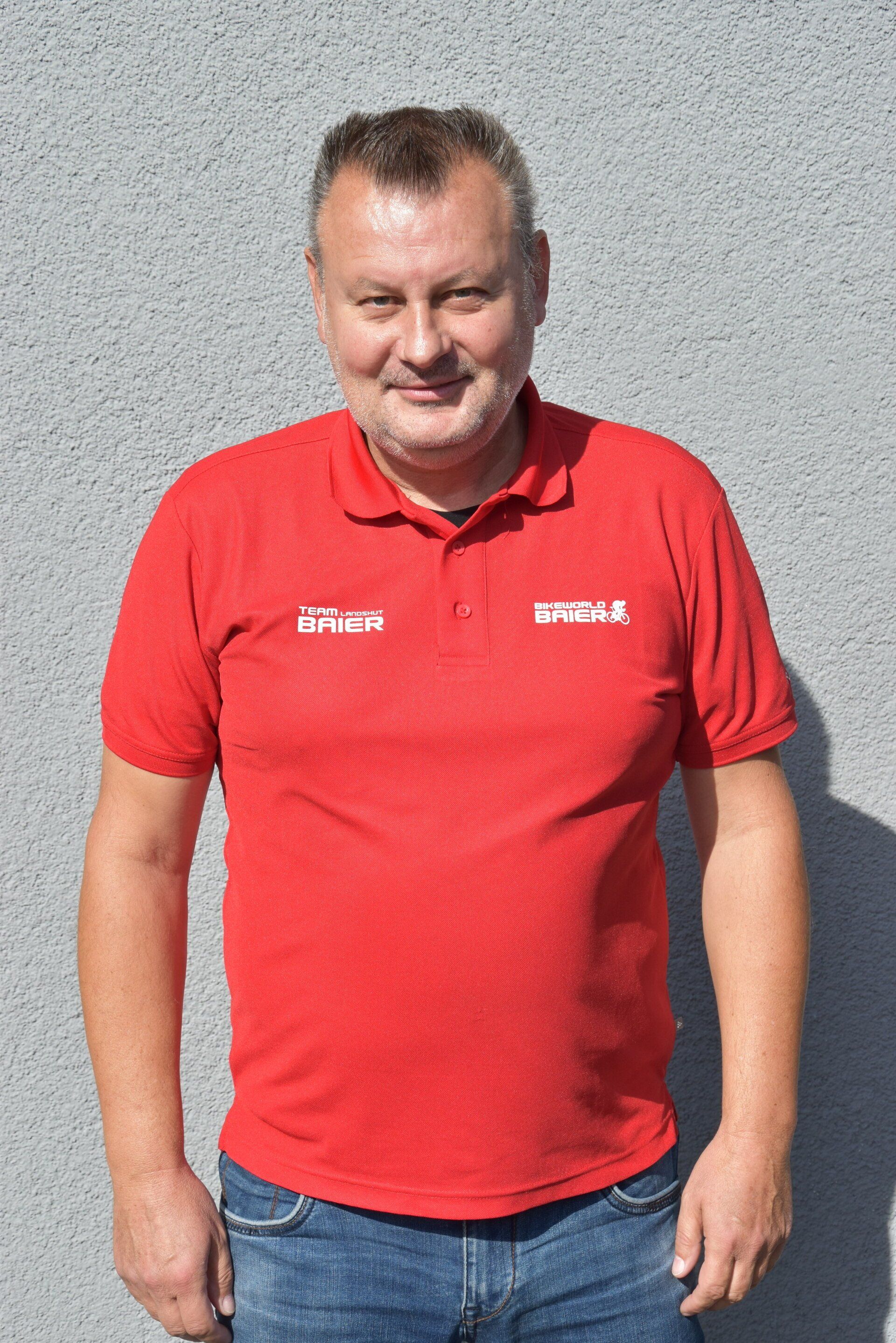 Ein Mann in einem roten Poloshirt und Jeans steht vor einer Wand.