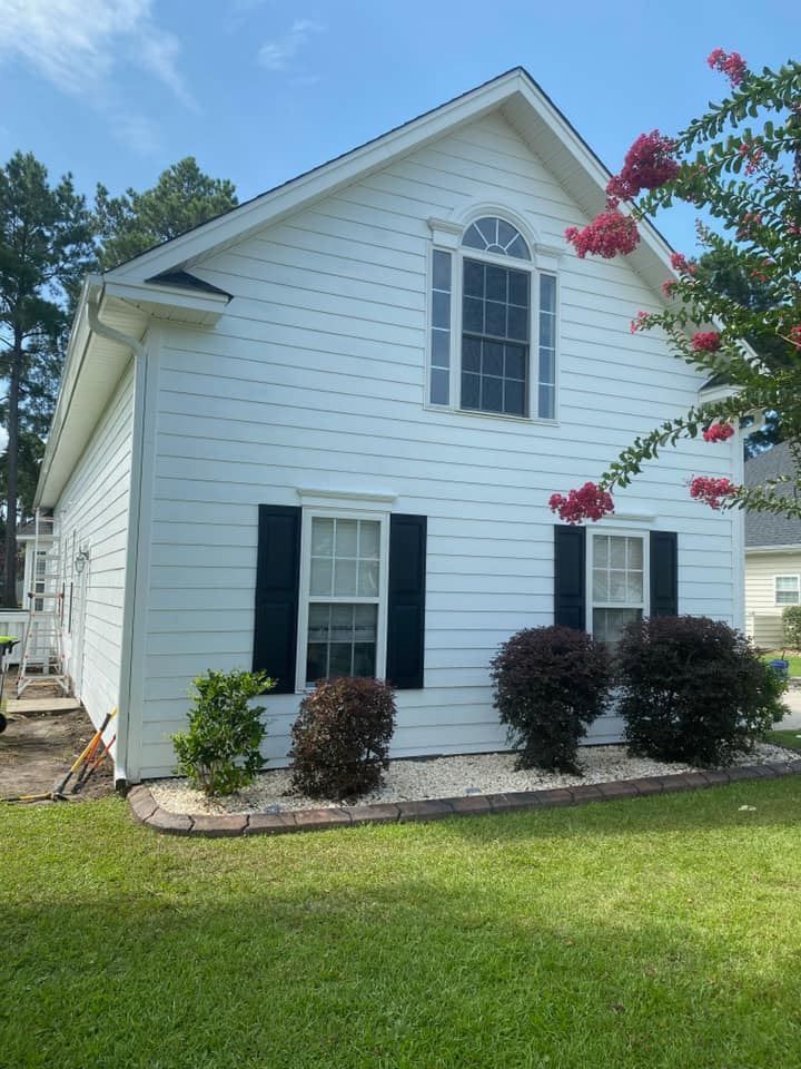 A white house with black shutters and flowers in front of it.
