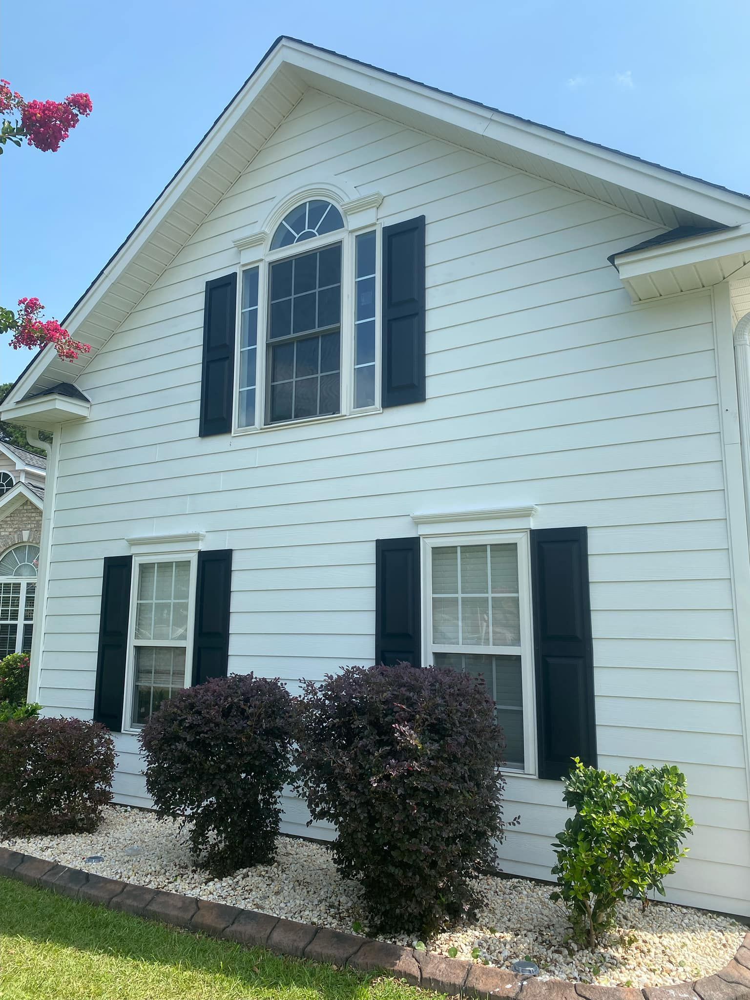 A white house with black shutters and bushes in front of it.