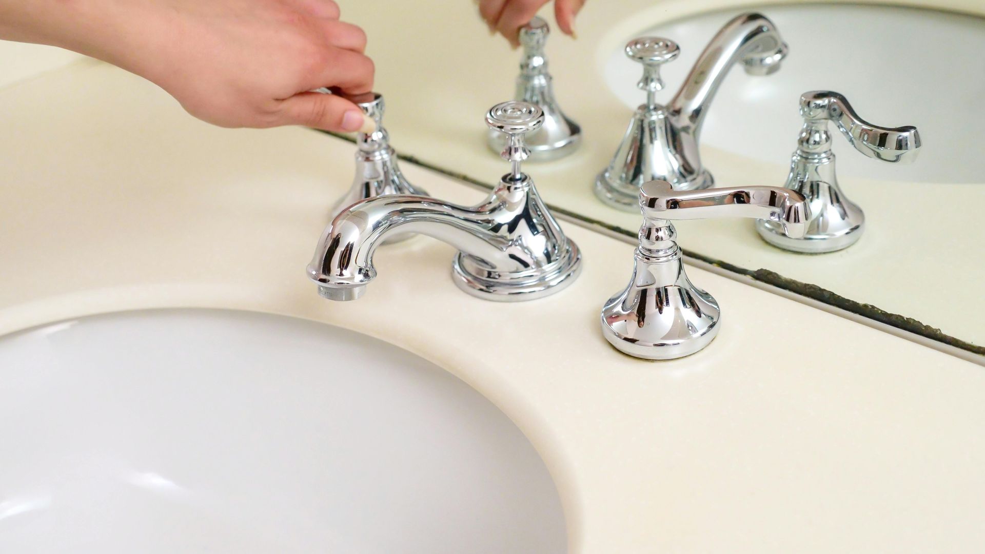 A hand adjusts a shiny chrome bathroom faucet in front of a vanity mirror.