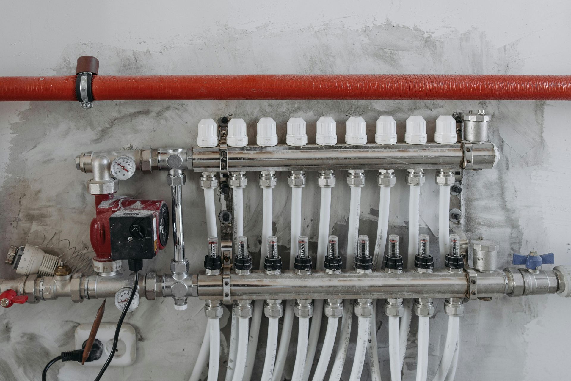 A hydronic radiant floor heating manifold system with a circulating pump, pressure gauges, and white tubing mounted on a wall.