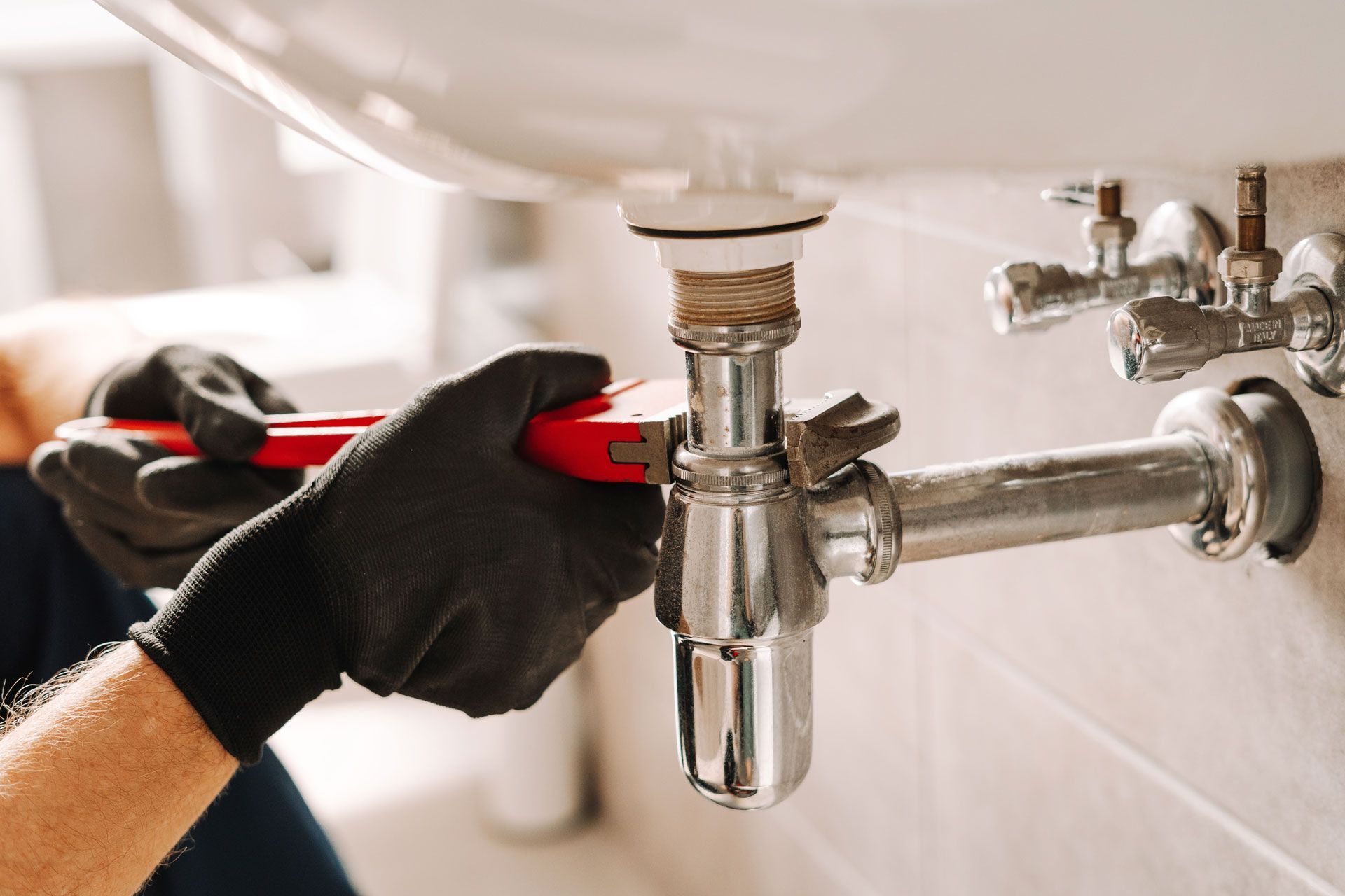 A person with black gloves uses a red wrench to tighten a pipe under a white sink in a bathroom.