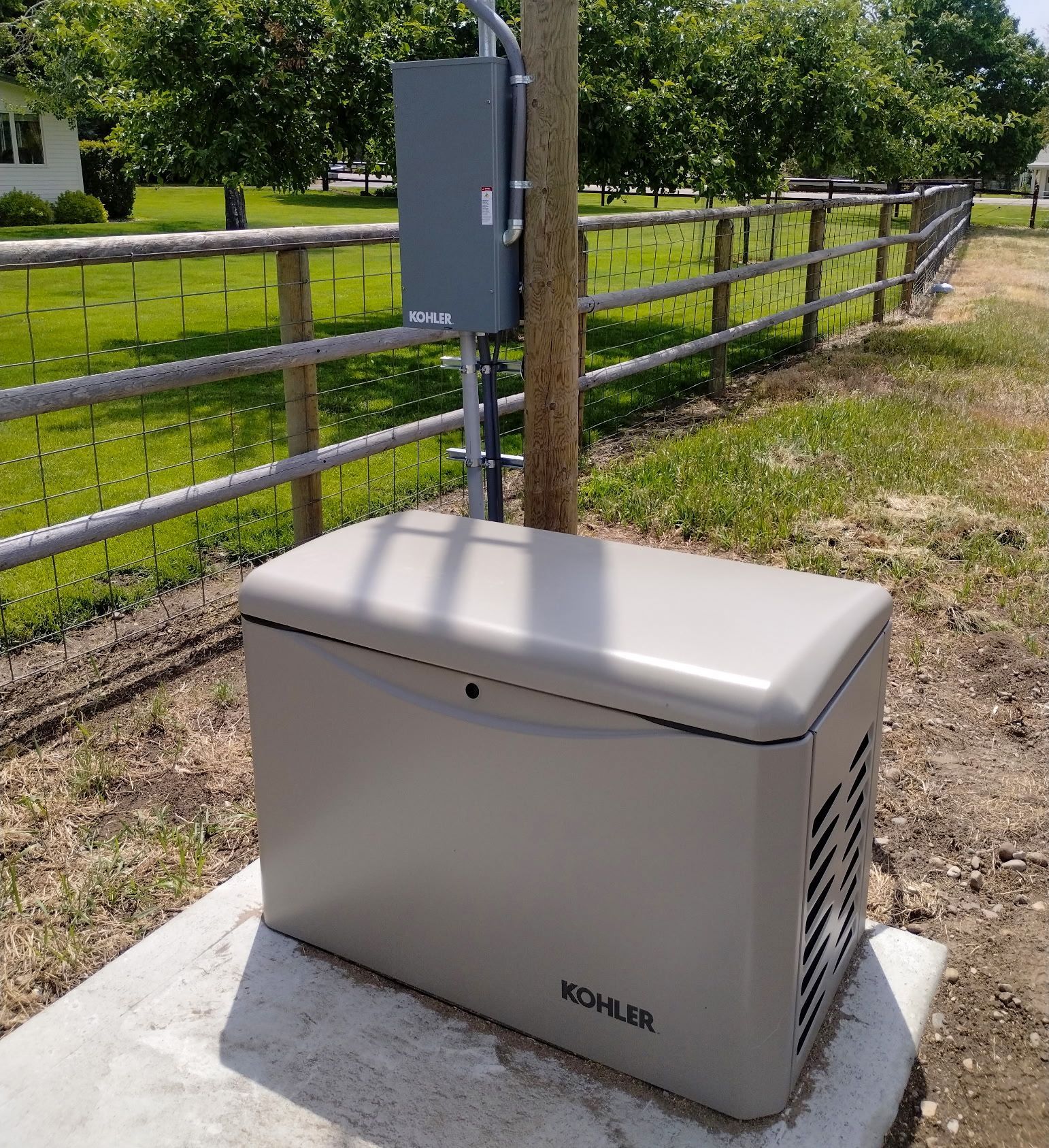 Kohler generator on concrete pad next to a pole with electrical box, near a fence and grass.
