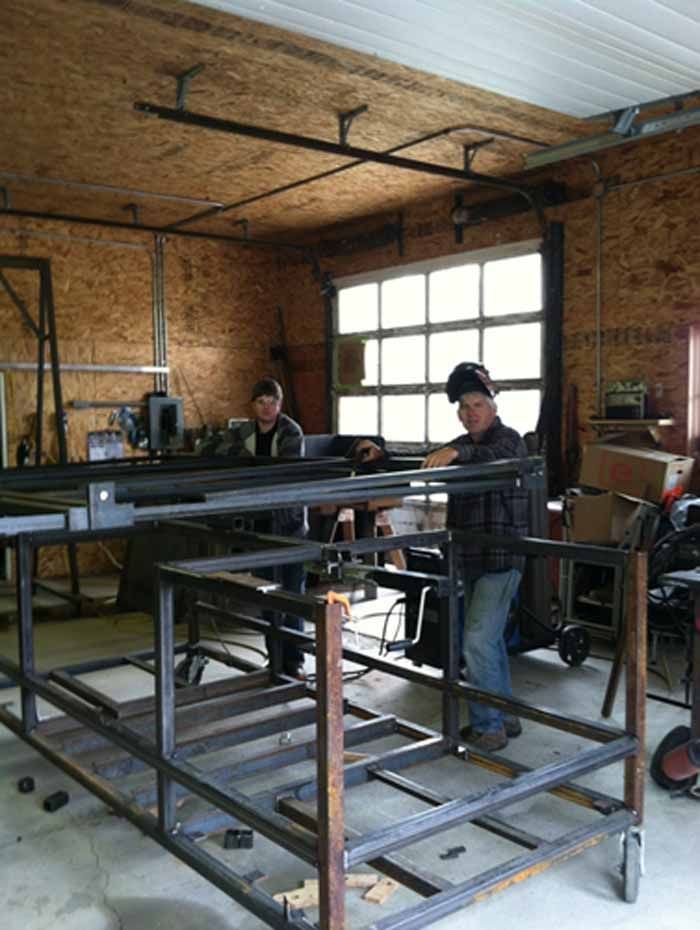 Two people welding metal frame inside a workshop; large window in background.